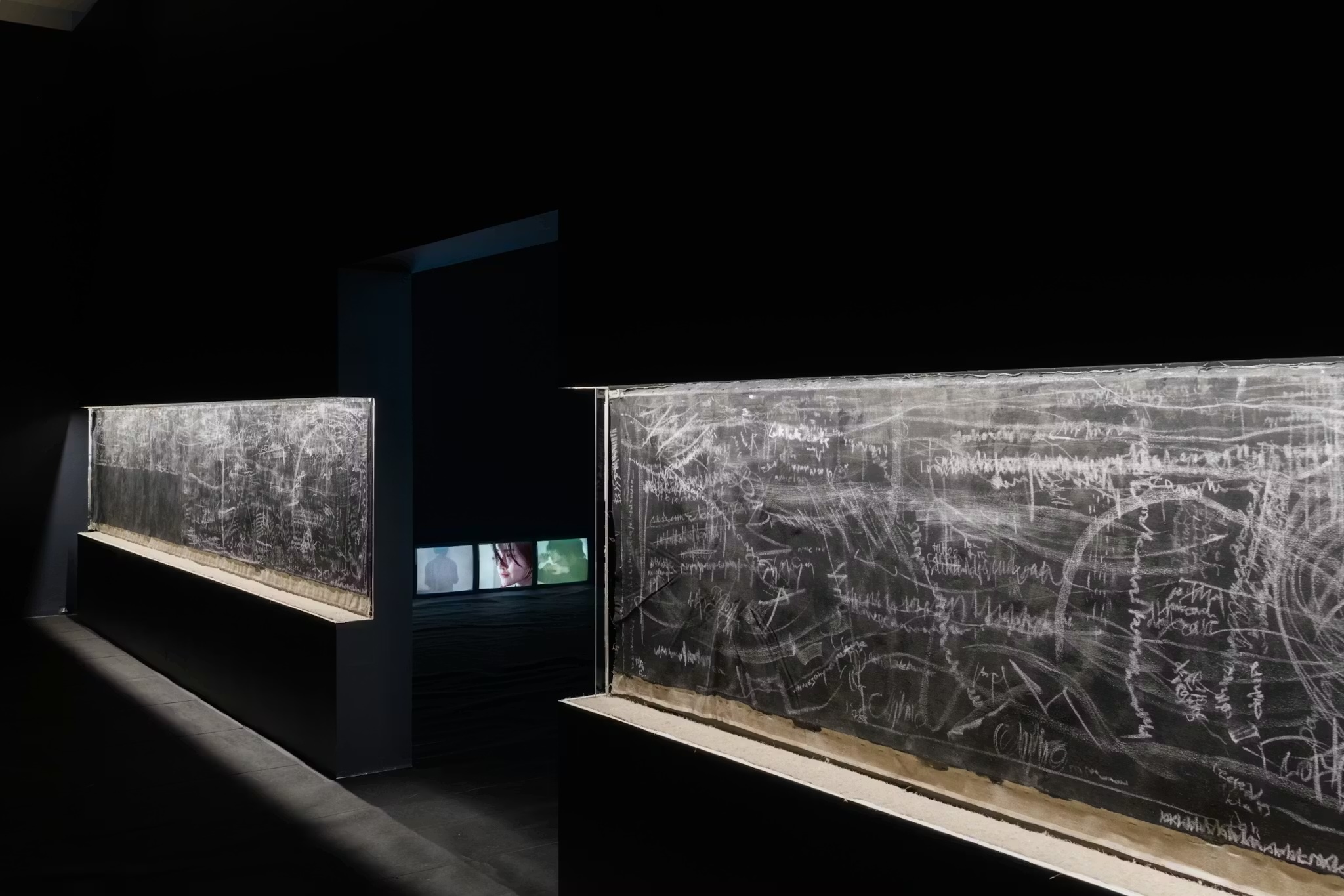 Installation by Pan Daijing. At the entrance to the exhibition space are seen two walls, each with a deep recess housing a black and white drawing. Each drawing is reminiscent of a busy chalkboard, with white writing, streaks, scribbles, and erasings seen atop a black background. Each drawing is also illuminated by a strip light at the top of the recess. Through the entrance between the two walls is visible a series of 3 televisions on the floor, each playing a different video piece by the artist. The first displays a silhouette drenched in a soft blue light. The second displays a close up of a persons face with their hair hanging over their eyes. The third displays a figure with their hands in front of their face drenched in soft green light. The entire exhibition space, with the exception of the artworks themselves, is pitch black.