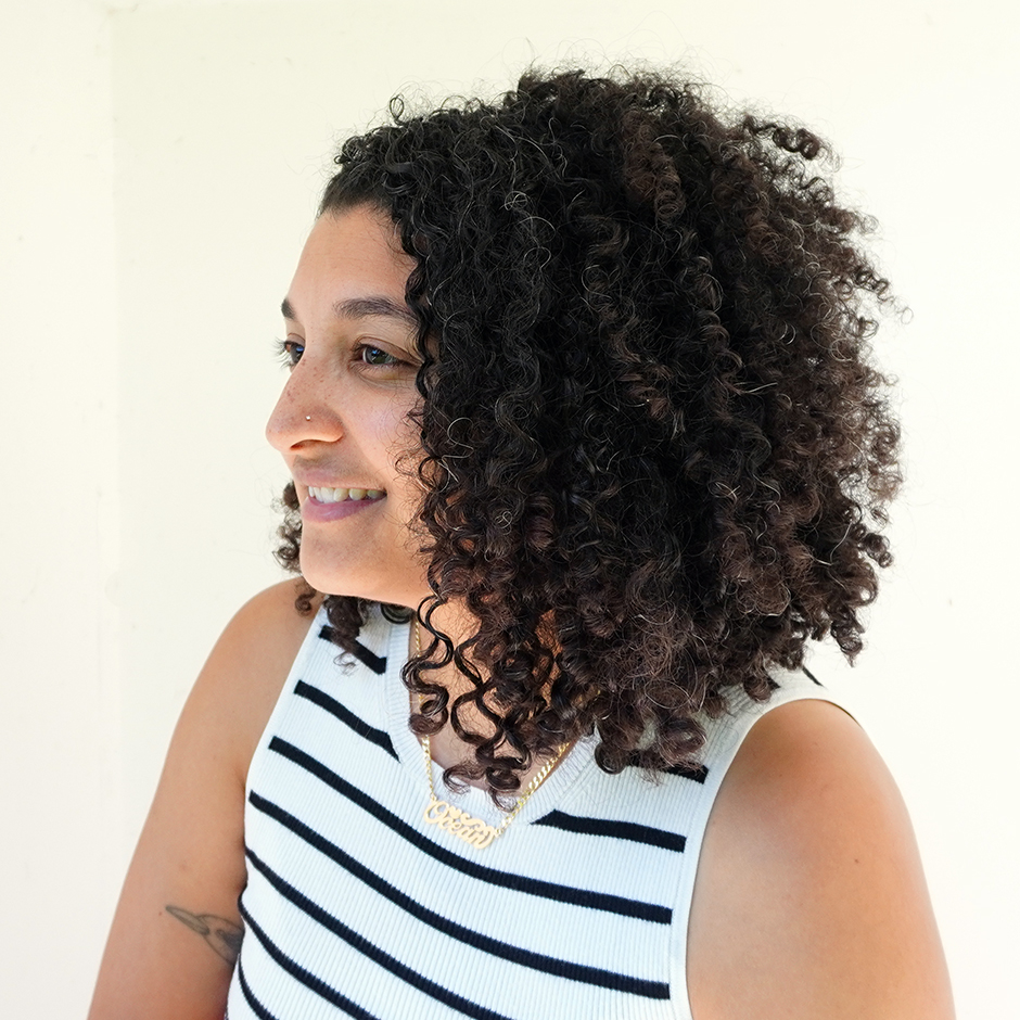 Profile portrait of a smiling sonia louise davis dressed in a black and white striped sleeveless shirt. Her curly hair is cropped into an asymmetrical bob. She is positioned in front of a white wall. 