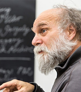 Profile portrait of Joseph Grigely dressed in a gray jacket with white hair and beard.