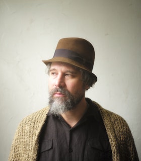 Portrait of Joshua Abrams dressed in a black button up underneath a beige wool cardigan, a beige dress hat and gray beard. He body faces forward as his face is turned slightly on the side.  