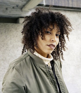 Portrait of Ligia Lewis looking up at the camera, with curly brown hair, red lipstick, and a green bomber jacket.