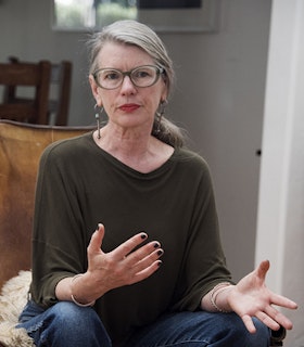 Portrait of Lisa Robertson sitting on a chair wearing a dark green sweater and blue glasses with her long grey hair back in a ponytail. Her hands are mid-motion as though she is orating.