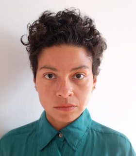 Mariana Valencia stares into the camera with short, curly, dark hair and a teal button up shirt against a white wall.