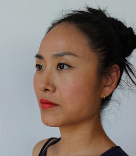 Portrait of Mika Tajima slightly side ways to the viewer with a messy top knot, black eyeliner, vibrant red lipstick and a black tank top.