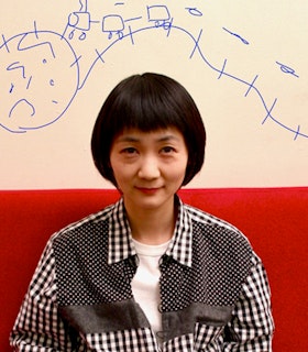 Portrait of Mina Nishimura sitted on a red surface and dressed in a patterned black and white button up with a white shirt underneath. The artist's hair is styled in a short bob with bangs and on the wall above the head a doodle drawn in blue.