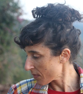 Portrait of Netta Yerushalmy with their hair up in a bun, looking down sideways and wearing a plaid yellow, blue, and red shirt.