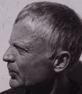 A close up, profile black and white portrait of Robert Grenier against a white textured wall. He has short hair and wears a dark shirt. 