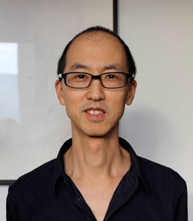 A portrait of Tan Lin against a shiny reflective white wall. He has short black hair and wears black glasses and a black shirt. 
