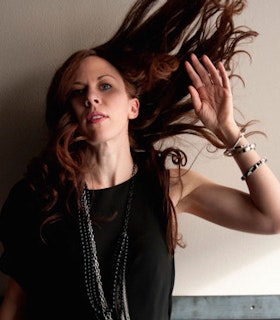 Portrait of Missy Mazzoli flicking her long red hair up in the air, arm raised, and wearing a black top.