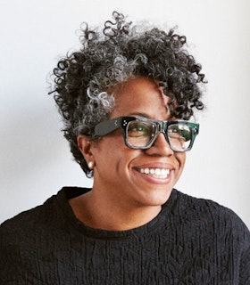 Portrait of Jennie C. Jones smiling in front of a white wall, wearing thick black glasses and a black sweater.