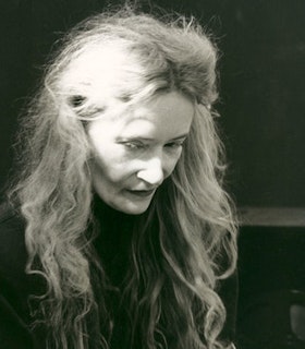 A close up black and white photographof Maryanne Amacher in front of a dark background. She has long light hair and wears a dark top. She looks downwards.