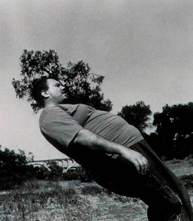 A black and white tilted portrait of Martin Kersels in a grassy place. He wears a t-shirt and dark pants and looks downwards. He has short hair. 