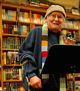 A portrait of David Meltzer performing in front of a wall of bookshelves. He wears a blue sweater, multicolored knit hat, a multicolored scarf, and thin circular glasses. In front of him, there is a music stand and a microphone.  