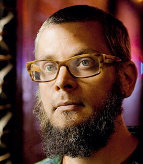 Close up portrait of Nate Wooley, with a curly dark beard and wearing tortoise-colored rectangular glasses.
