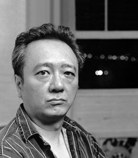 A black and white portrait of John Yau looking intently at the camera. He wears a striped shirt and is in front of an opened window.