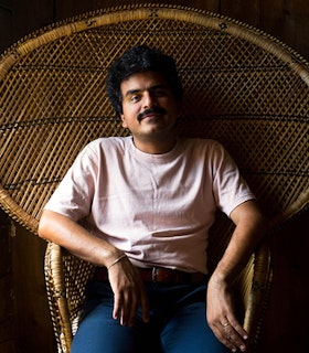 Portrait of Roberto Carlos Lange dressed in a pink shirt, with blue jeans and a brown belt. Adorning a mustache and black curly hair the artist leans down on a weaved chair with a flared back design.
