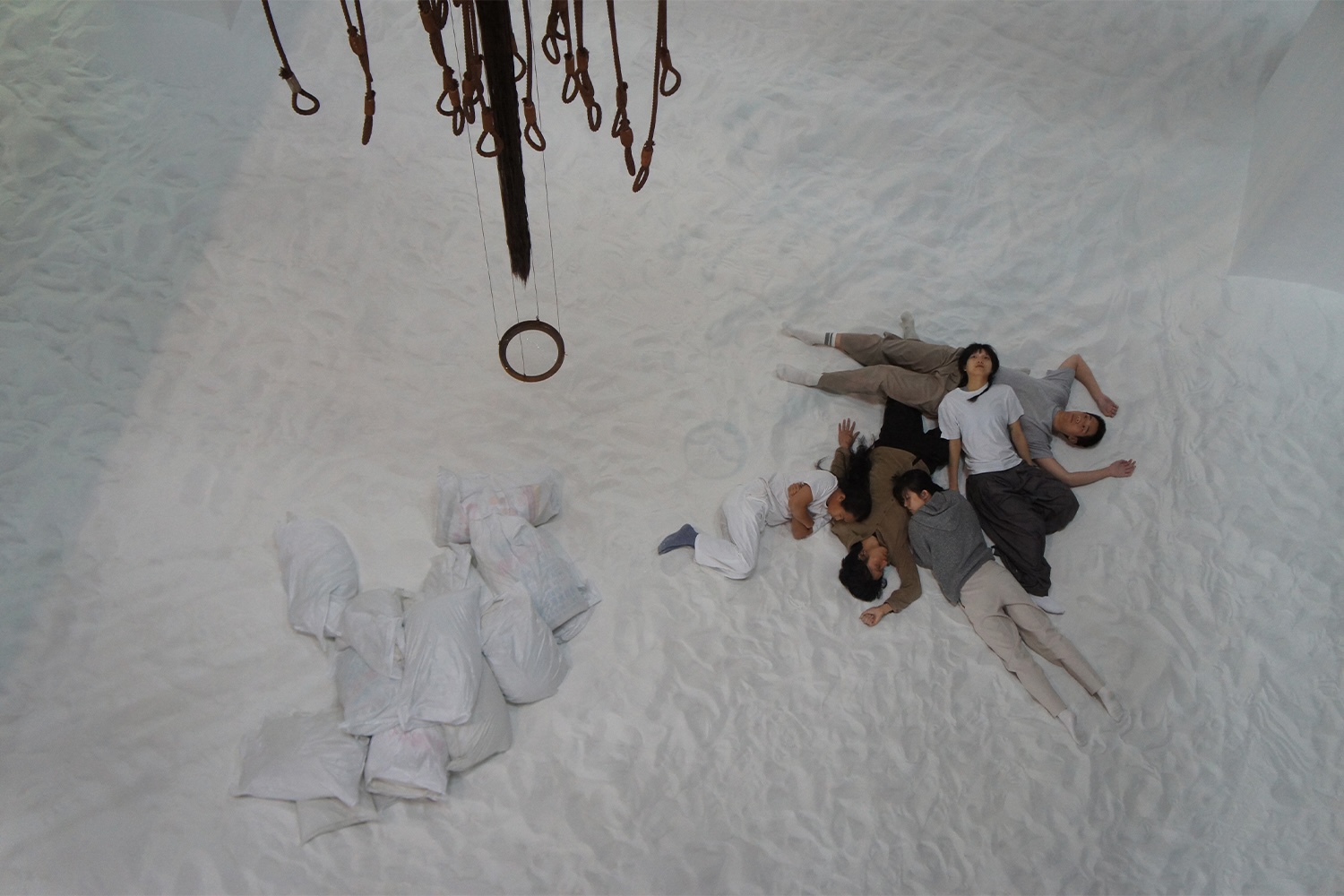 A group of performers are seen lying on the floor within an installation by Pan Daijing. The performers are seen wearing sweatpants, sweatshirts, and t-shirts in a variety of muted colors including white, grey, brown, and taupe. They are lying in a pile on top of one another on a floor entirely covered in white sand. To the left of the performers are seen a small pile of white garbage bags. Above the performer hang a mobile-like sculpture created using rusted metal cables.