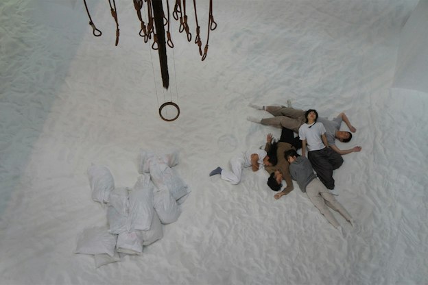 A group of performers are seen lying on the floor within an installation by Pan Daijing. The performers are seen wearing sweatpants, sweatshirts, and t-shirts in a variety of muted colors including white, grey, brown, and taupe. They are lying in a pile on top of one another on a floor entirely covered in white sand. To the left of the performers are seen a small pile of white garbage bags. Above the performer hang a mobile-like sculpture created using rusted metal cables.