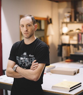 A portrait of Sam Lewitt in front of a a work table with arms crossed, wearing a black graphic T-shirt.