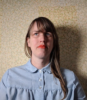Portrait of Sharah Hennies in front of a yellow wallpaper with flowers, dressed in a blue shirt with orange lipstick and blond straight hair with bangs swept to one side.