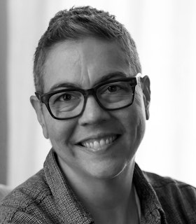 Grayscale prortrait of a smiling Stacy Szymaszek with a pixie haircut, button up shirt and thick framed glasses.