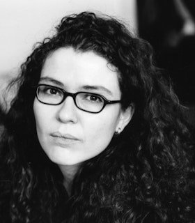 Black and white portrait of Sylvie Courvoisier wearing dark framed glasses and long dark curly hair that fall on her front.