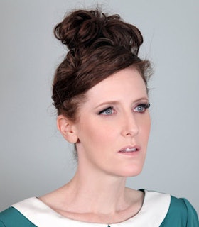 Portrait of Tina Satter staring into the distance, with her hair up in a bun and wearing a green top with a white collar in front of a grey wall.
