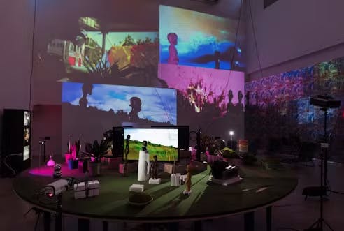 Round green table with various small statues, pots, books, white clay and a still screen of a grassy area scattered around surrounded by cameras. Behind it a projection of four wall-size images depicting the statues as silhouettes in front of grassy areas and plants.