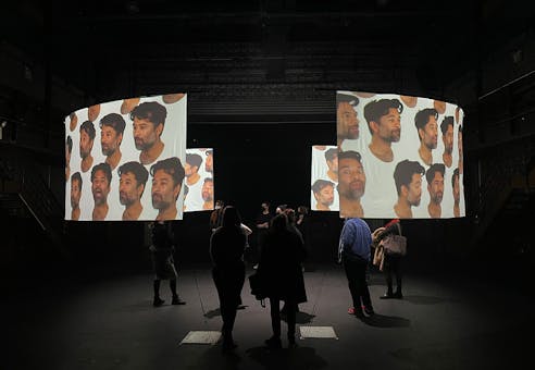 A dark gallery space with two curved projection screens showing images of the same person with varying expressions. Several audience members are viewing the screens from various vantage points.