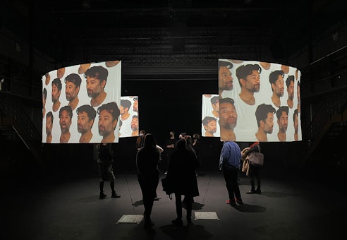 A dark gallery space with two curved projection screens showing images of the same person with varying expressions. Several audience members are viewing the screens from various vantage points. 