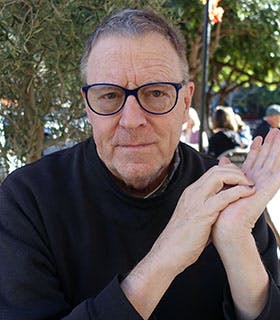 George Albon is centered and directly faces the camera. He is in sharp focus against a soft outdoor backdrop with greenery, wearing a dark sweater and blue-framed glasses, his hands clasped together in front of him.