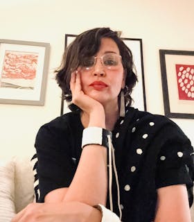 Layli Long-Soldier sits on a couch looking straight ahead. She gazes down at the camera, with her chin resting on one hand. She wears a black, patterned shirt, white cuff on each arm, long earlings, and glasses. In the background, there is framed artwork against a beige wall.