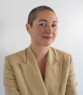 Jacqueline Kiyomi Gork is slightly angled toward the camera, looking directly into the lens. They are wearing a beige blazer against a clean, softly lit white background.
