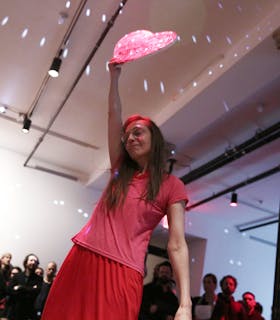 Jutta Koether is photographed mid-performance in an indoor space. She is wearing a pink top and a red skirt, holding a glowing pink object high above her head. A group of people observe from the background.
