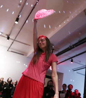 Jutta Koether is photographed mid-performance in an indoor space. She is wearing a pink top and a red skirt, holding a glowing pink object high above her head. A group of people observe from the background.