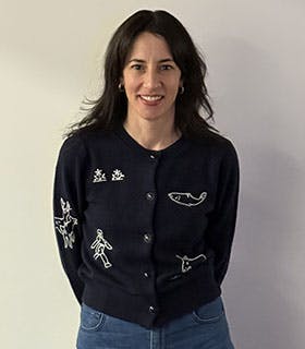 Jennifer Lafferty is centered, smiling directly at the camera. She stands straight against a light, neutral wall, wearing an embroidered navy cardigan, hoop earrings, and jeans.