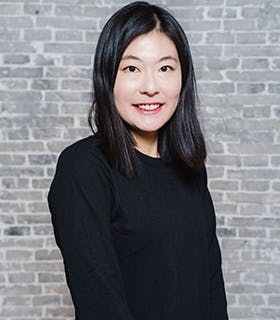 Fi Jae Lee stands before a slightly blurred brick background under bright lighting. She smiles, facing the camera with her body angled slightly to the right. She wears a black long-sleeve shirt with her hands at her sides.