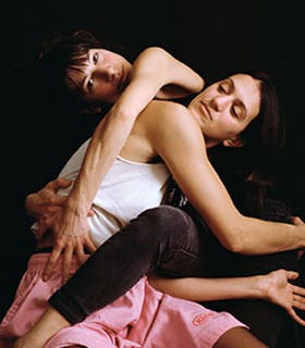 Lisa Fagan and Lena Engelstein are photographed intertwined against a black background. Lisa is positioned in the back, her head tilted to the left with her arms around Lena’s torso. Lena is in the foreground, seated and gazing to the left, with her arms embracing Lisa’s legs. Lisa is wearing all black, while Lena is in a white tank top and pink shorts.