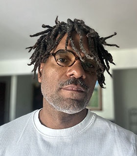 Javier Cardona Otero looks directly at the camera with his head tilted slightly. He is standing indoors against a softly lit background, and wearing round brown glasses and a white textured shirt.