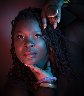 Ebony Noelle Golden looks directly into the camera, resting her chin on one hand, with the other hand extended over her head in a dramatic pose. She is illuminated by vibrant red and blue lighting against a dark backdrop, and is wearing a nose ring, several bracelets, and a large ring.