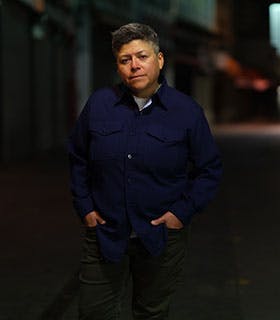 Raquel Gutiérrez is centered and directly faces the camera, standing outdoors at night in a softly lit urban setting. She wears a navy button-up shirt and green pants, her hands casually in her pockets. The background is blurred, with subtle hints of storefronts and streetlights.