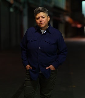 Raquel Gutiérrez is centered and directly faces the camera, standing outdoors at night in a softly lit urban setting. She wears a navy button-up shirt and green pants, her hands casually in her pockets. The background is blurred, with subtle hints of storefronts and streetlights.