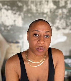 Jennifer Harge looks directly at the camera in a close-up portrait. She is wearing a black tank top and layered gold necklaces, set against a textured wall with a workspace and art supplies visible in the background.