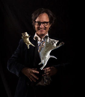 Asher Hartman grins at the camera as two kittens appear to leap from his body towards the foreground. He is wearing rectangular-framed glasses, a dark suit, and a patterned tie, and stands in front of a black background.