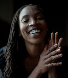 Shayla-Vie Jenkins is smiling against a black background with her hands raised in front of her body and clasped together. She is wearing a dark top under soft, neutral lighting.