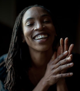 Shayla-Vie Jenkins is smiling against a black background with her hands raised in front of her body and clasped together. She is wearing a dark top under soft, neutral lighting.