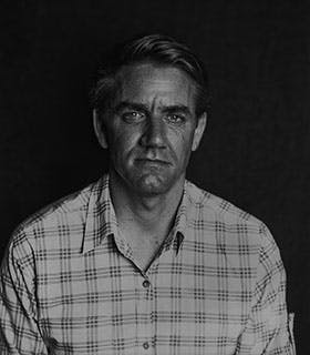A black and white portrait of Jon Kinzel, who is centered and staring straight at the camera. Against a dark background, he is in high contrast, wearing a light plaid shirt.