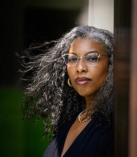 Khadijah Queen gazes at the camera, her body turned to the right, her hair blows with the wind. She wears pointed glasses, bright jewelry, and a navy blouse, and appears in front of a blurred background with greenery.