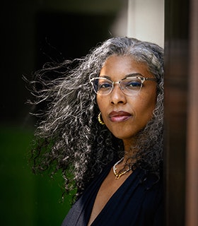Khadijah Queen gazes at the camera, her body turned to the right, her hair blows with the wind. She wears pointed glasses, bright jewelry, and a navy blouse, and appears in front of a blurred background with greenery. 