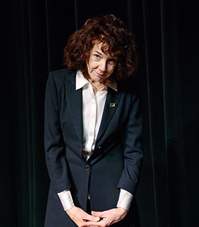 Mary Ruefle gazes up at the camera, shrugging her shoulders. She wears a navy blue suit and a white button down. Her hands clasped, she stands against green velvet curtains under a bright light. 